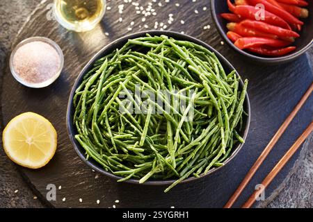 Frische Salicornia Algen in einer schwarzen Schüssel mit Zitrone, Sesamsamen, Chili-Paprika und Essstäbchen, Draufsicht Stockfoto