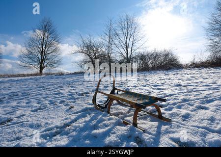Ein hölzerner Schlitten sitzt auf einem schneebedeckten Hang unter einem klaren blauen Himmel mit hellem Sonnenschein. Fußabdrücke sind im Schnee zu sehen, was auf kürzliche Aktivitäten hinweist Stockfoto