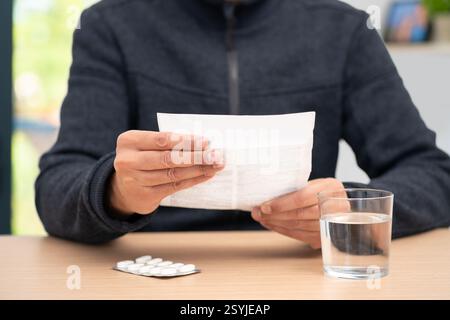 Schuss eines nicht erkennbaren Mannes Hände, die ein Faltblatt halten und am Tisch in seinem Haus lesen, mit einer Tablette Pillen und einem Glas Wasser Stockfoto