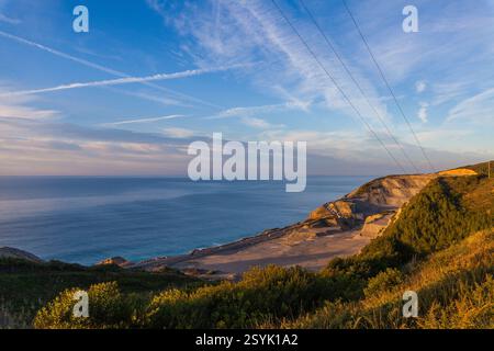 Eine Küstenlandschaft erstreckt sich bis zum Horizont, mit einer felsigen Klippe mit einem Steinbruch- oder Ausgrabungsort, mit Blick auf das weite Meer unter blauem Himmel Stockfoto
