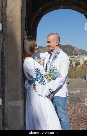 Sommerhochzeit in Georgien: Romantisches Paar in ukrainischen bestickten Outfits küsst sich unter einem alten Bogen Stockfoto