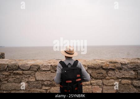 Senor Mann Tourist mit Hut, der das Meer in der Nähe einer Steinmauer an bewölktem Herbsttag sieht. Rentenreisekonzept Stockfoto