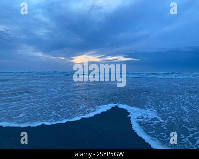 Zwei Wellen verschmelzen über nassem Sand an einem Winterstrand, während das letzte goldene Licht der Sonne durch die Wolken am fernen Horizont bricht. Stockfoto