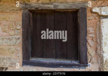 Ein dunkler, verwitterter Holzladen bedeckt eine Fensteröffnung in einer rauen Steinmauer. Stockfoto