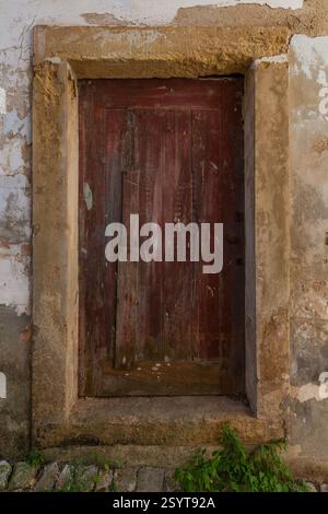 Eine verwitterte, dunkelrote Holztür mit einem Steinrahmen ist in eine zerbröckelnde Wand eingefasst, an der Unkraut wächst Stockfoto