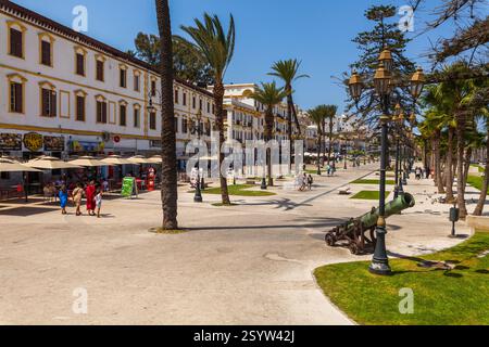 Lebhafte mediterrane Promenade gesäumt von Palmen, Cafés und historischen Gebäuden, mit einer Kanone im Vordergrund und Menschen, die entlang der W spazieren Stockfoto
