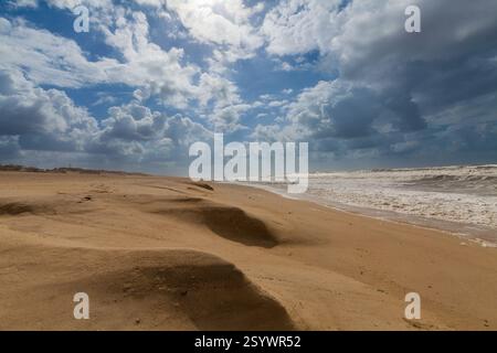Ein riesiger Sandstrand erstreckt sich in Richtung eines turbulenten Ozeans unter einem dramatischen Himmel voller flauschiger weißer Wolken und blauer Flecken. Stockfoto