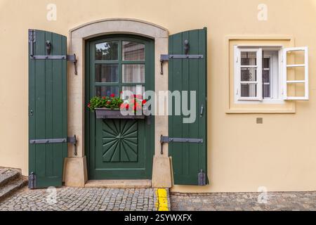 Die charmante Hausfassade verfügt über eine lebendige grüne Tür mit dekorativen Schnitzereien, passenden Fensterläden, einem Fensterkasten mit roten Blumen und einem angrenzenden weiß Stockfoto