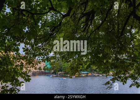 Eingerahmt von üppigen grünen Ästen entfaltet sich eine Flussszene mit Booten, die über das Wasser verstreut sind, Gebäuden am fernen Ufer und einem bewölkten Himmel darüber. Stockfoto