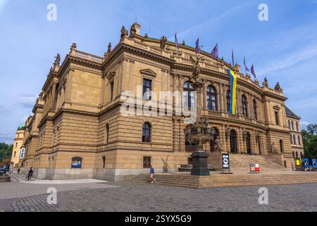 Ein großes, reich verziertes Gebäude mit Bogenfenstern, dekorativen Statuen und Fahnen, darunter ein langes gelbes und blaues Banner, steht in einem gepflasterten Quadrat Stockfoto