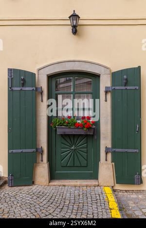 Die charmante Hausfassade verfügt über eine lebendige grüne Tür mit dekorativen Schnitzereien, passenden Fensterläden, einem Fensterkasten mit roten Blumen und einem angrenzenden weiß Stockfoto