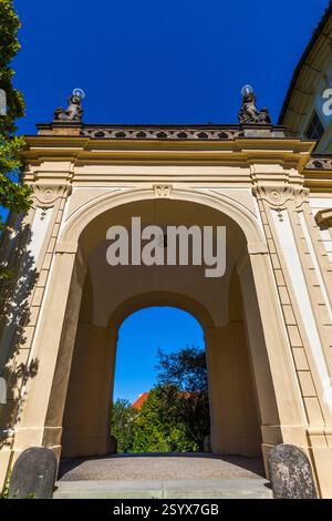 Ein flaches Bild von einem bogenförmigen Tor mit dekorativen Statuen oben, die einen Blick auf Bäume und Gebäude vor einem hellblauen Himmel bieten. Stockfoto