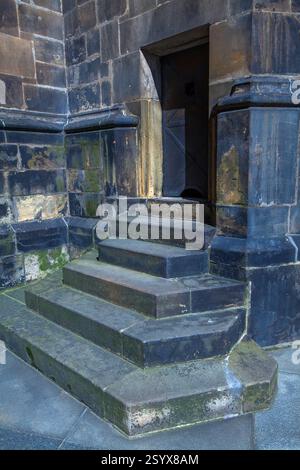 Eine Nahaufnahme der verwitterten Steintreppen führt zu einer dunklen Eisentür, die in einer dicken Steinmauer mit alten Texturen, moosigen Oberflächen und starkem GE untergebracht ist Stockfoto