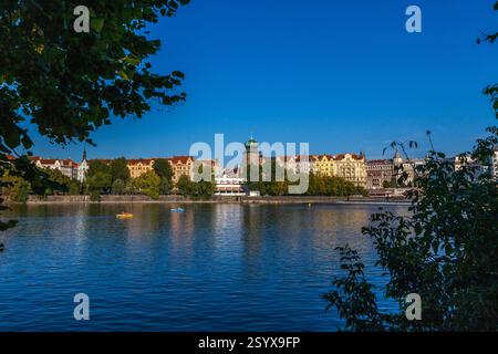 Ein breiter Fluss, der den blauen Himmel reflektiert, mit bunten Gebäuden am Ufer, Bäumen im Vordergrund und kleinen Booten, die auf dem Wasser liegen. Stockfoto