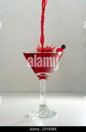 Vorderansicht des Stroms von rotem Likör, der in Kirschcocktail in konischem Glas auf Keramiktisch vor grauem Hintergrund spritzt Stockfoto
