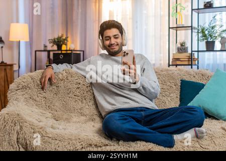 Junger indischer Mann auf dem Sofa, der kabellose Kopfhörer trägt Musik auf dem Smartphone hört wählt den Lieblingstitel entspannt und tanzt. Arabian Guy genießt Rhythmus, der sich mit Energie und fröhlichem Ausdruck bewegt. Stockfoto