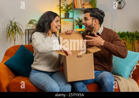 Glückliches, aufgeregtes Paar, das zusammen auf dem Sofa sitzt. Kunden aus hispanischen jungen Familien packen ein gutes Paket mit Blick nach innen aus und bieten fünf großartige Einkäufe per Postversand Stockfoto