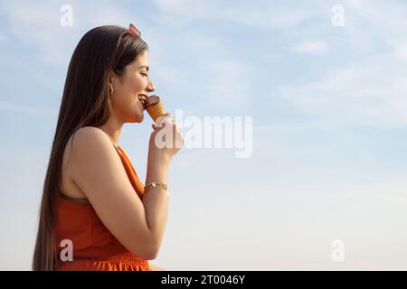 Porträt einer jungen Frau, die Eis im Kegel vor blauem Himmel isst Stockfoto