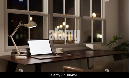 Seitenansicht eines modernen Büroarbeitsplatzes bei Nacht mit einem Laptop-Modell mit weißem Bildschirm, einer Tischlampe und Zubehör auf einem Tisch vor dem Fenster. Stockfoto