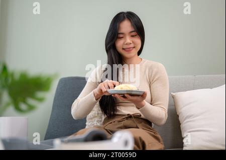 Eine schöne und fröhliche junge asiatische Frau genießt ihr Frühstück und isst gebackenes Brot auf einem Sofa in ihrem Wohnzimmer. Menschen- und Lebensmittelkonzepte Stockfoto