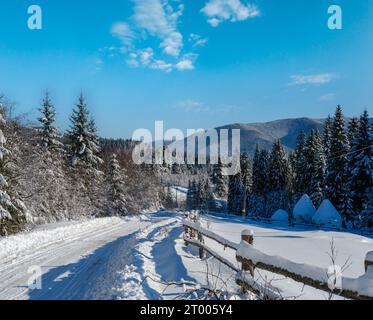 Sekundäre Landschaft alpine Straße zu abgelegenen Bergdörfern durch verschneiten Tannenwald, Schneeverwehungen und Holzzaun auf dem Weg Stockfoto