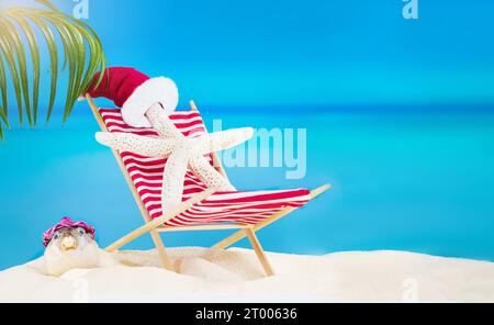 Weißer Seestern mit Weihnachtsmann-Hut auf gestreifter roter Sonnenliege mit Kugelfischen am Sandstrand mit Palmen hinter dem Meer. Weihnachten, Neujahr, Sommer, Trav Stockfoto