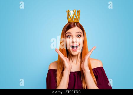 Eine Frau mit dem überraschten Gesicht und der Krone auf dem Kopf öffnete den Mund und die Augen weit und hielt die Hände in der Nähe ihrer Wangen, isoliert auf gelbem Hintergrund Stockfoto