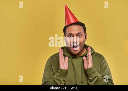 Ein wunderbarer junger indianer mit Geburtstagshut und khakifarbenem Kapuzenpullover, mit den Händen näher am Gesicht, Geburtstag Stockfoto