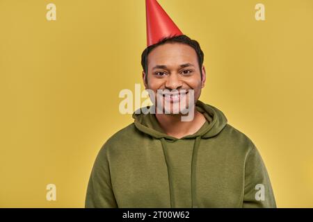 Fröhlicher junger indischer Mann in Geburtstagshut und lässigem Khaki Hoodie lächelt in die Kamera, Geburtstag Stockfoto