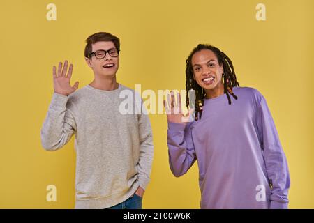 Zwei fröhliche Freunde in lila und weißen Sweatshirts, die vor der Kamera winken und lächeln, Vielfalt Stockfoto