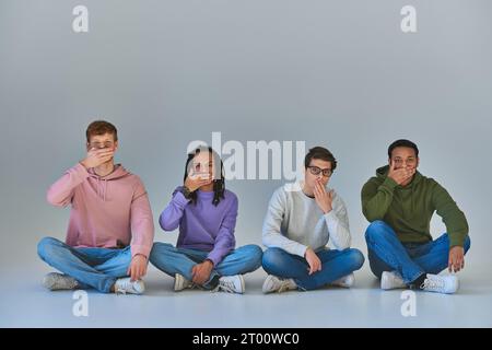 Vier multikulturelle Freunde sitzen mit überkreuzten Beinen und bedeckenden Mündern, kulturelle Vielfalt Stockfoto