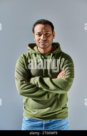 Junger indischer Mann im khakifarbenen Hoodie lächelt mit gekreuzten Händen auf der Brust auf gelbem Hintergrund Stockfoto