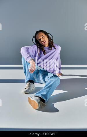 Hübscher afroamerikanischer Mann im Sweatshirt mit Dreadlocks, der auf dem Boden sitzt und in die Kamera blickt Stockfoto