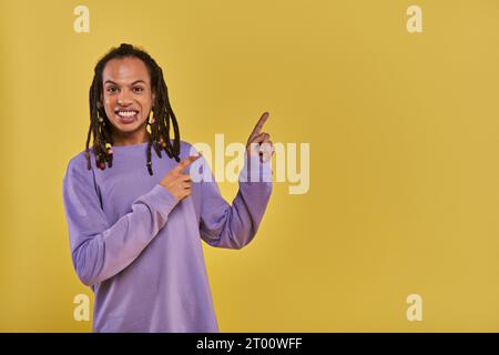 Fröhlicher afroamerikanischer Mann mit Dreadlocks, durchbohrter Lippe, die auf die rechte Seite zeigt, vor gelbem Hintergrund Stockfoto