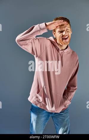 Fröhlicher junger Mann in trendigem Hoodie und Jeans, der Hand an der Stirn hält und vor grauem Hintergrund posiert Stockfoto