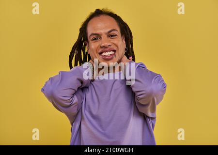 Fröhlicher afroamerikanischer Mann mit Dreadlocks und durchbohrter Lippe, der seinen Hals berührt und aufrichtig lächelt Stockfoto