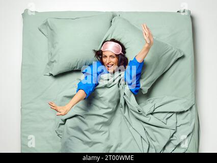 Junge schöne Frau mit Schlafmaske bedeckt mit Decke, die Arme zum Aufwachen ausdehnt. Genug Schlaf für die Nacht. Am frühen Morgen. Stockfoto