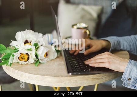 Frau Hände arbeiten am Laptop im Café. Nahaufnahme. Freiberufler mittleren Alters mit einer Tasse Kaffee Tippen und Lesen. Loft-Arbeitsplatz mit Blumen Stockfoto