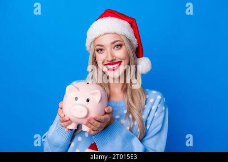 Foto-Porträt der attraktiven blonden jungen Frau halten Schweinchen Finanzier tragen Pullover x-MAS Hut isoliert auf blauem Hintergrund Stockfoto