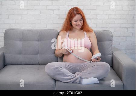 Rothaarige schwangere Frau sitzt auf der Couch und benutzt einen heimdoppler. Stockfoto