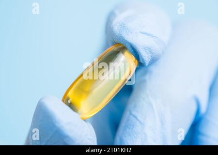 Arzt, Apotheker oder Wissenschaftler Hand in blauen Handschuhen mit Omega-3-Kapsel. Vitaminpille oder -Ergänzung auf blauem Hintergrund. Gesundheitswesen und Wissenschaft Stockfoto