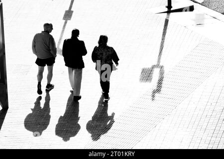 Drei Leute laufen die Straße runter. Das starke Licht hebt ihre Silhouetten und ihre Schatten auf dem Boden hervor. Stockfoto