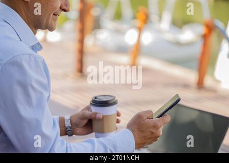 Detail der Hände eines lateinischen Mannes, der ein Handy und eine Einwegtasse mit Kaffee hält. Stockfoto