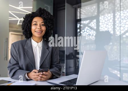 Porträt einer jungen, erfolgreichen afroamerikanischen Geschäftsfrau, die an einem Schreibtisch in einem Büro an einem Schreibtisch sitzt, selbstbewusst in die Kamera blickt und ein Telefon in der Hand hält. Stockfoto