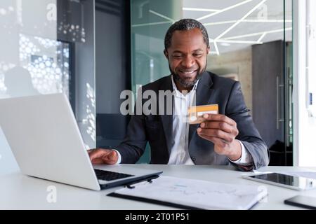 afroamerikaner lächelnder männlicher Geschäftsmann, der im Büro am Laptop arbeitet und Kreditkarte hält und benutzt. Stockfoto