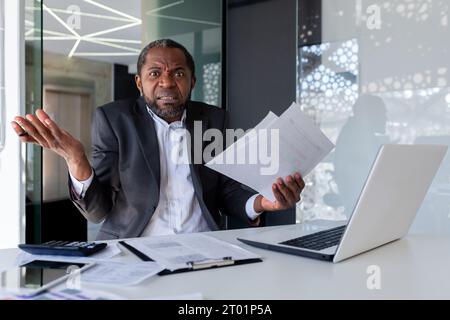 Porträt eines besorgten afroamerikanischen Mannes, der Dokumente, Papiere und Vereinbarungen in den Händen hält. Im Büro am Schreibtisch sitzen und frustriert mit den Armen in die Kamera werfen. Stockfoto