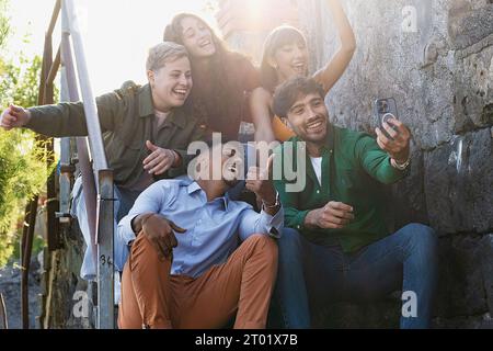 Eine Gruppe von fünf Personen, darunter eine nicht-binäre Person und ein afrikanischer Nachkomme, sind an der Treppe am Meer und fangen zusammen mit einem s ein freudiges Selfie ein Stockfoto