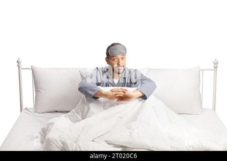 Nachdenklicher junger Mann im Pyjama, der in einem Bett auf weißem Hintergrund sitzt Stockfoto