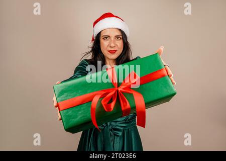 Studio-Aufnahme eines schönen glücklichen, freudigen, aufgeregten Mädchens in santa-Hut und grünem Kleid, das eine Geschenkbox in der Hand hält. Frohe Weihnachten. Neujahrskonzept Stockfoto