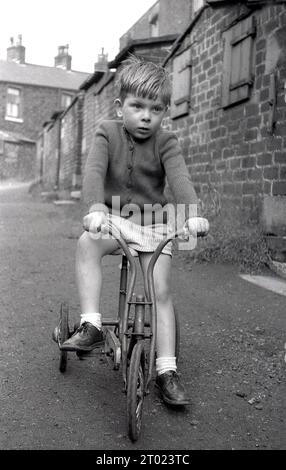 1960er Jahre, historisch, auf einer Schottergasse, ein kleiner Junge auf seinem dreirädrigen Metallroller, Oldham, England, Großbritannien. Stockfoto